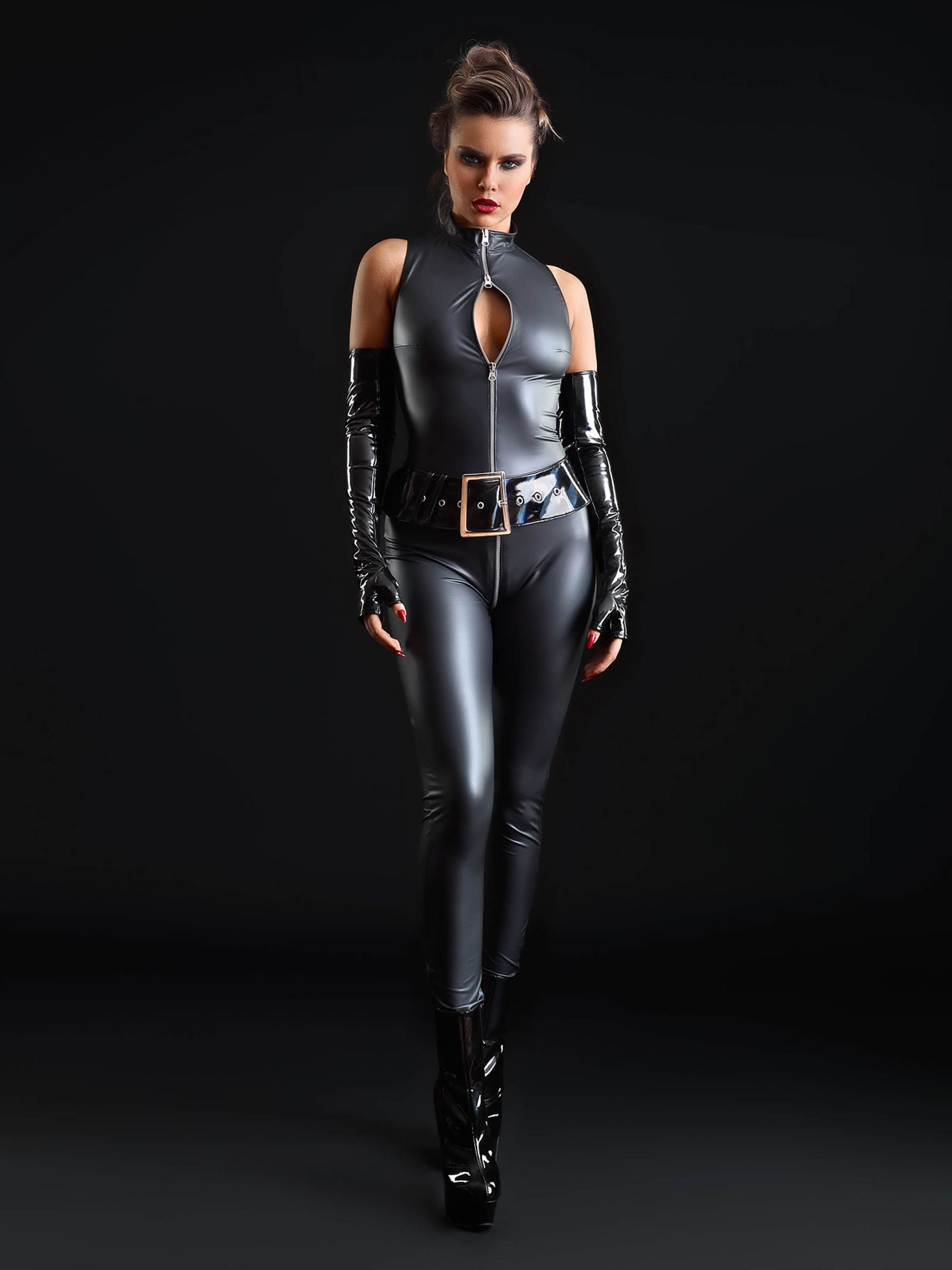 Frau posiert im ärmellosen Kunstleder-Catsuit mit Reißverschluss, Lackgürtel und Plateau-Heels vor schwarzem Hintergrund.
